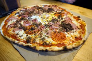 A freshly baked pizza topped with slices of ham, ground beef, and scattered herbs, resting on a piece of brown paper.

