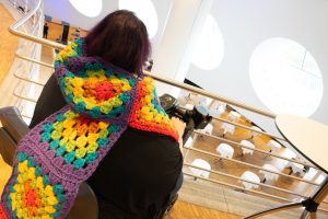 A rainbow scarf trails from a wheelchair as its wearer gazes out over a sunlit hall of circles and stories waiting to unfold.
