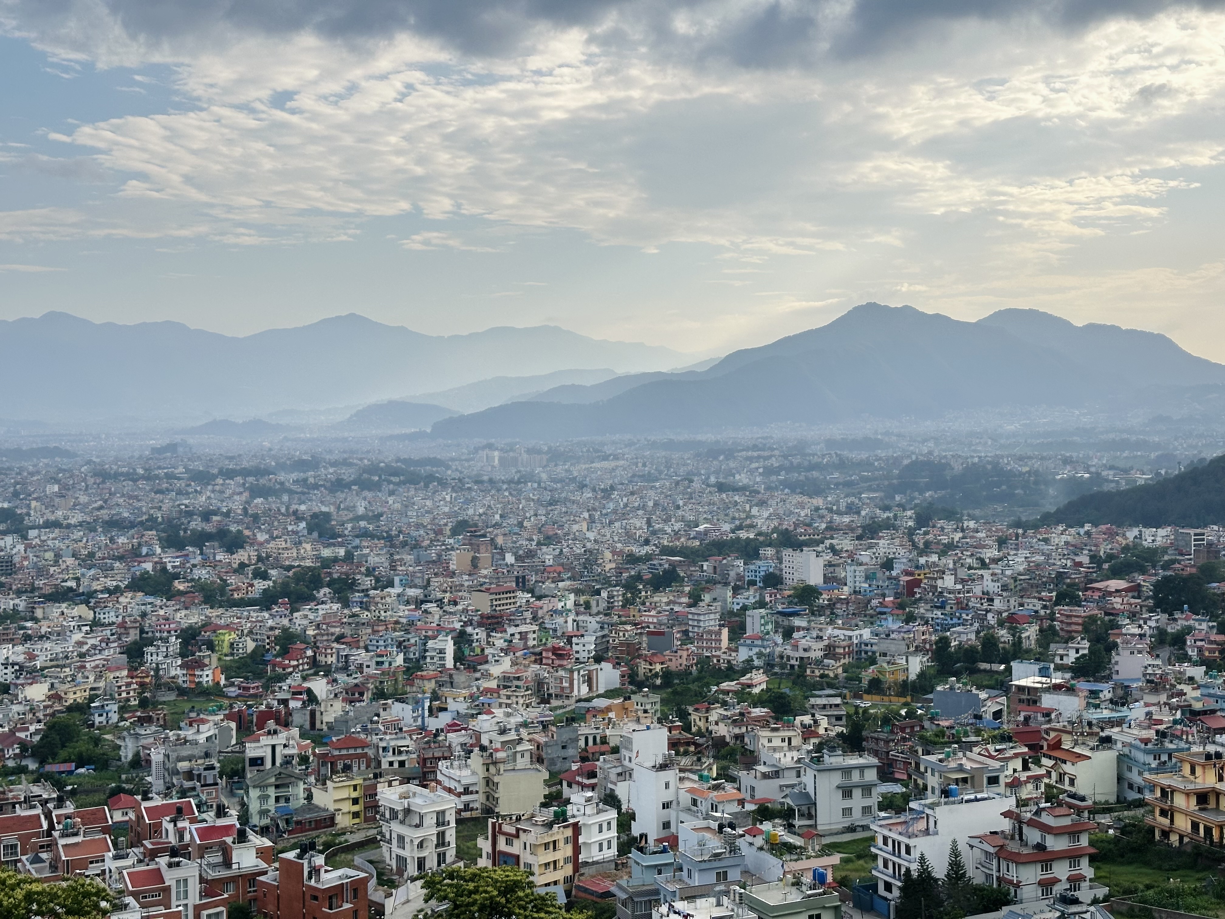 A panoramic view of a densely city surrounded by mountains under a cloudy sky.