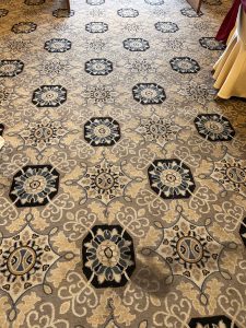 A patterned carpet featuring a repeated design of hexagons and flowers in shades of beige, black, and blue, with intricate swirling motifs. 