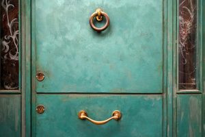 A weathered turquoise door with a round, bronze door knocker at the top and a horizontal brass handle near the bottom. Two small round locks are visible on the left side of the door, and there are two glass panels on either side, featuring intricate swirling designs. 
