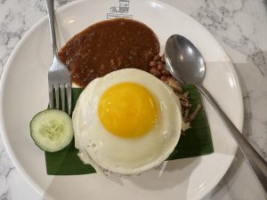 A plated meal featuring a fried egg with a runny yolk, placed on top of a small portion of rice, garnished with a green banana leaf.