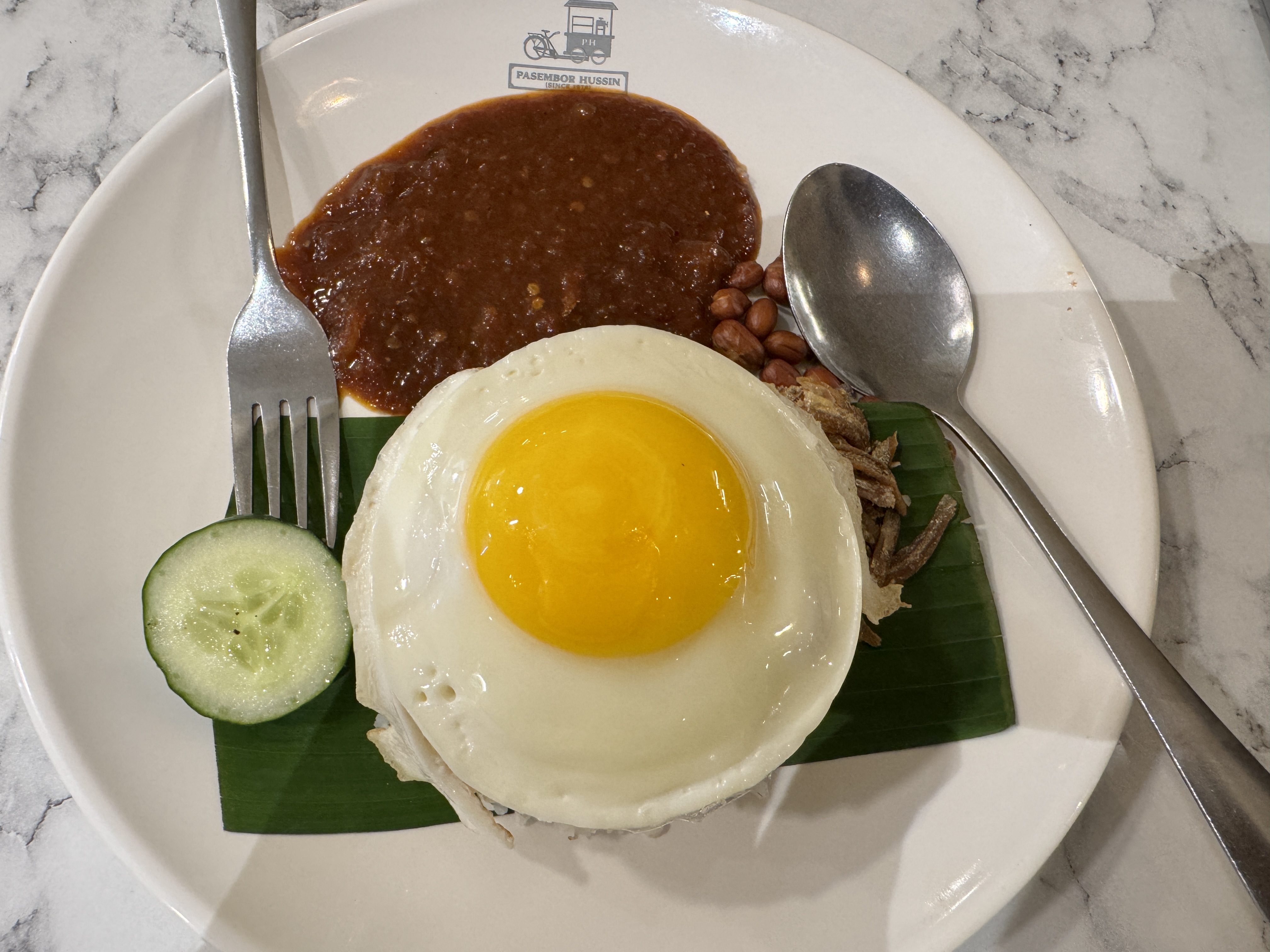 A plated meal featuring a fried egg with a runny yolk, placed on top of a small portion of rice, garnished with a green banana leaf.