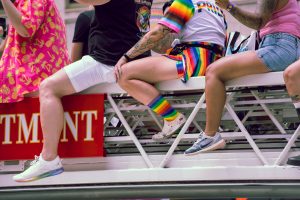 People dressed in colorful Pride attire sit on the ladder of a fire department truck during a parade. One person wears rainbow shorts, socks, and a “PRIDE” shirt.
