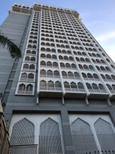 A tall, modern building with a façade featuring numerous arched windows, intricately designed white lattice work, and a gray concrete structure. The photo is taken from a low angle, emphasizing the height of the building against a clear blue sky.
