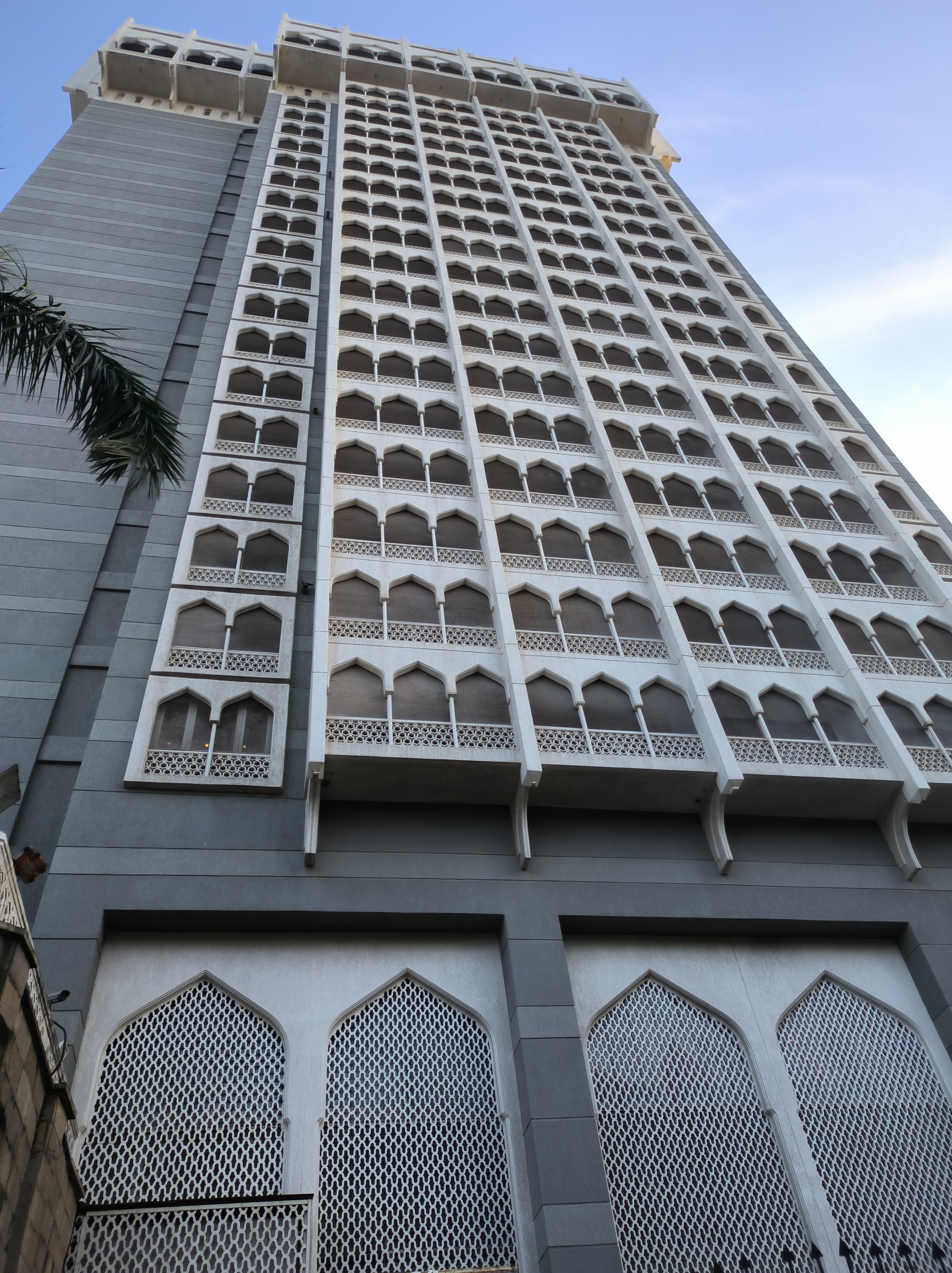 A tall, modern building with a façade featuring numerous arched windows, intricately designed white lattice work, and a gray concrete structure. The photo is taken from a low angle, emphasizing the height of the building against a clear blue sky.