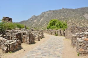 Bhangarh Fort's stone ruins in Rajasthan feature a paved path through crumbling structures, set against a barren hill.