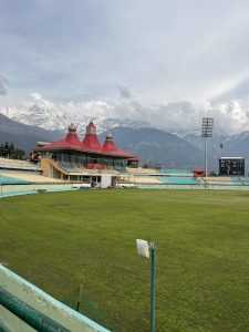 The photo shows a vibrant cricket stadium with a well-maintained green field. In the background, there is a red-roofed pavilion with domed towers. Snow-capped mountains rise majestically behind the stadium under a cloudy sky, creating a striking contrast with the lush green field.
