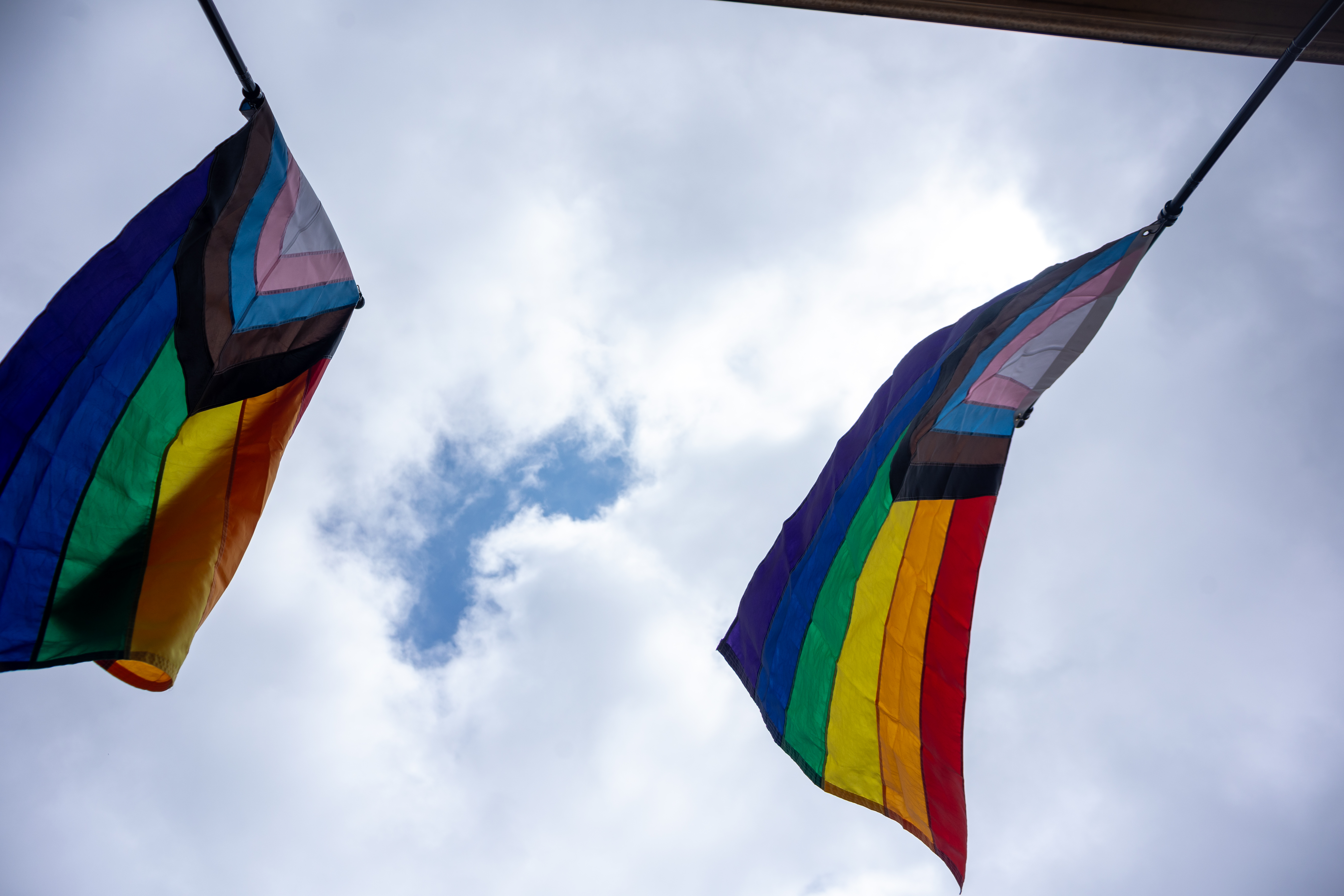 Looking up at two progress pride flags flying in the wind with just a hint of blue sky peaking out from the clouds