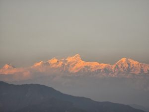 Snow-capped mountains bathed in a warm golden light at sunrise, with a soft mist in the foreground and rolling hills in silhouette below.