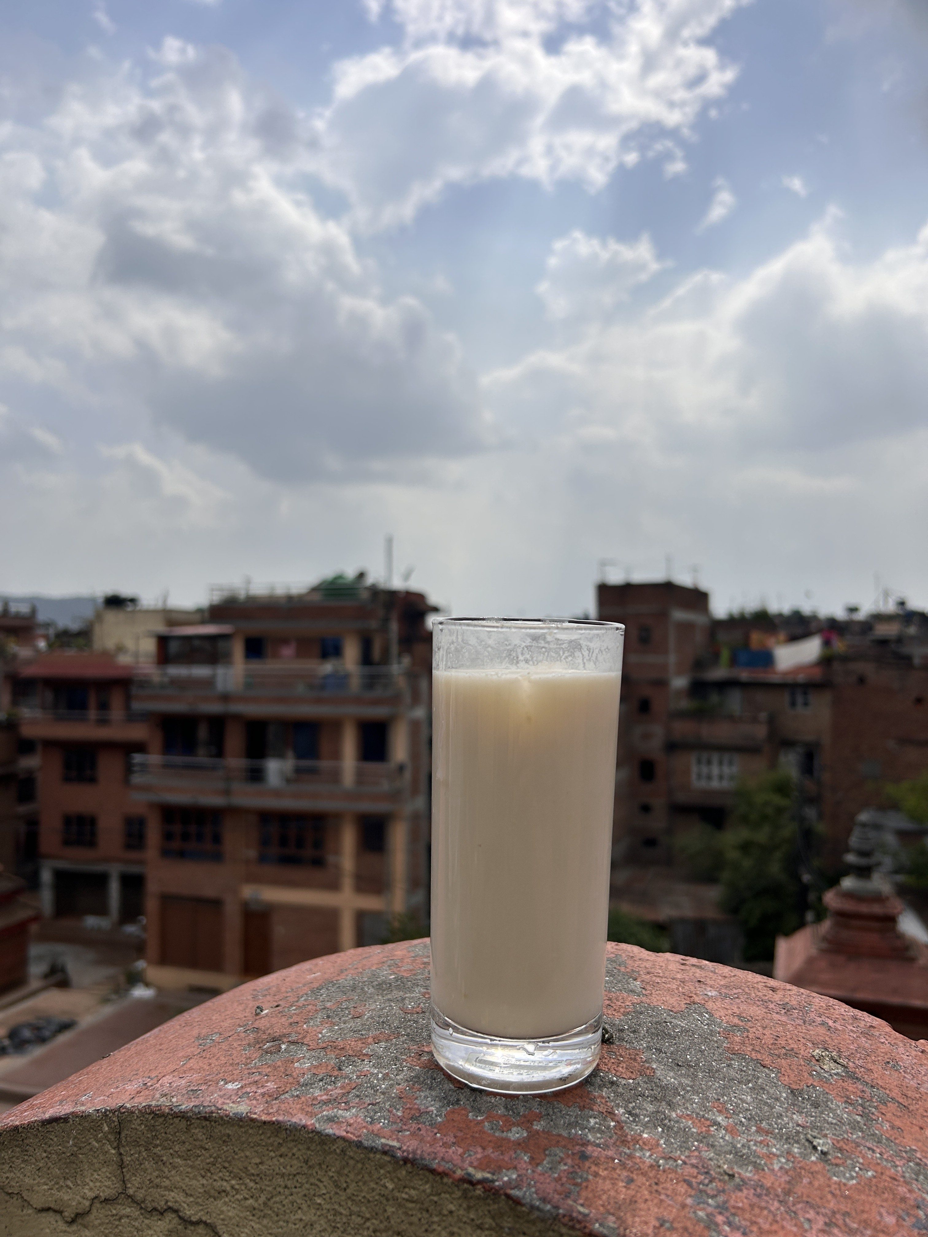 A glass of light-colored beverage is placed on a textured ledge, with a blurred urban landscape in the background featuring various buildings and a cloudy sky.