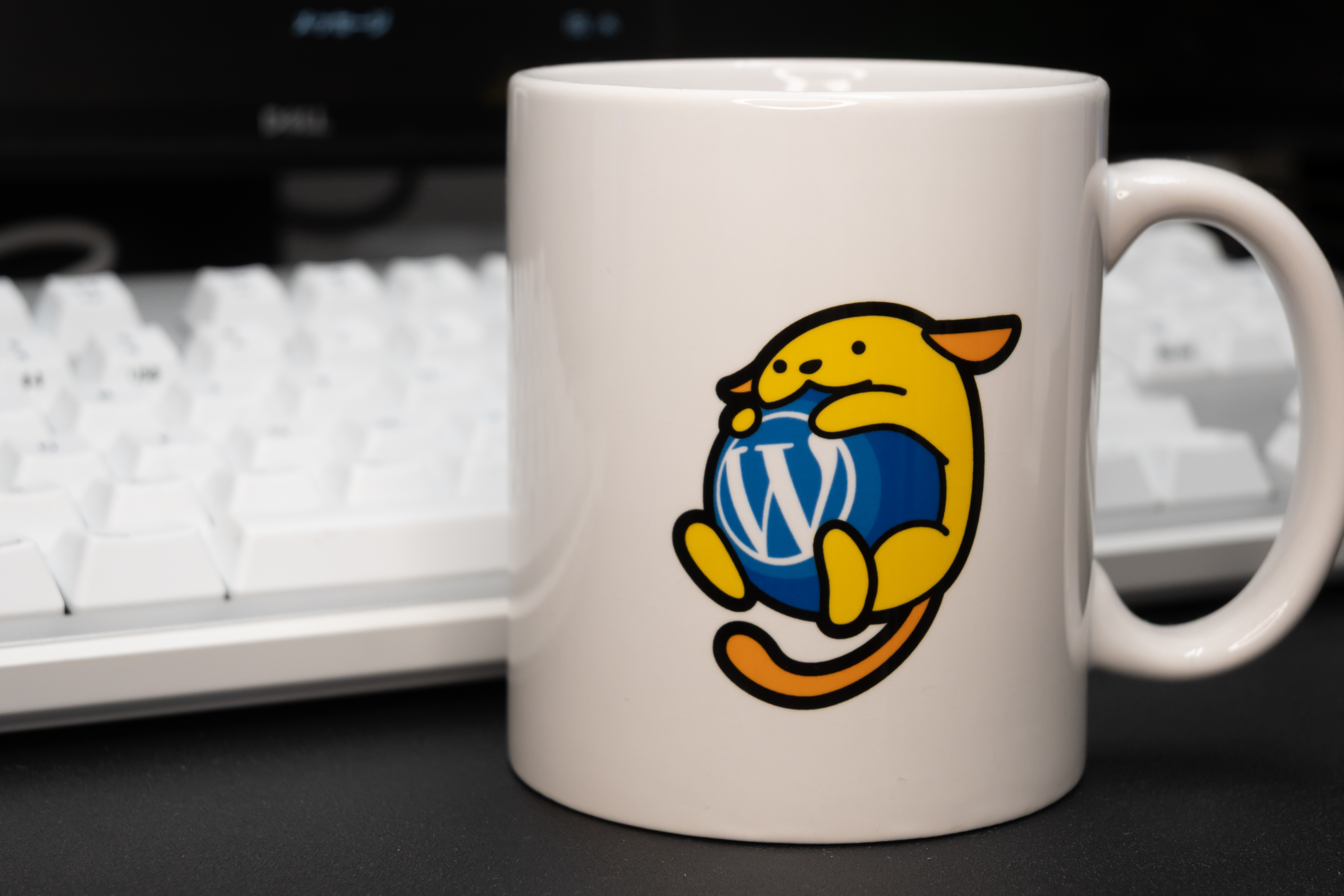 A white mug featuring Wapuu hugging the WordPress logo, placed in front of a keyboard on a desk.