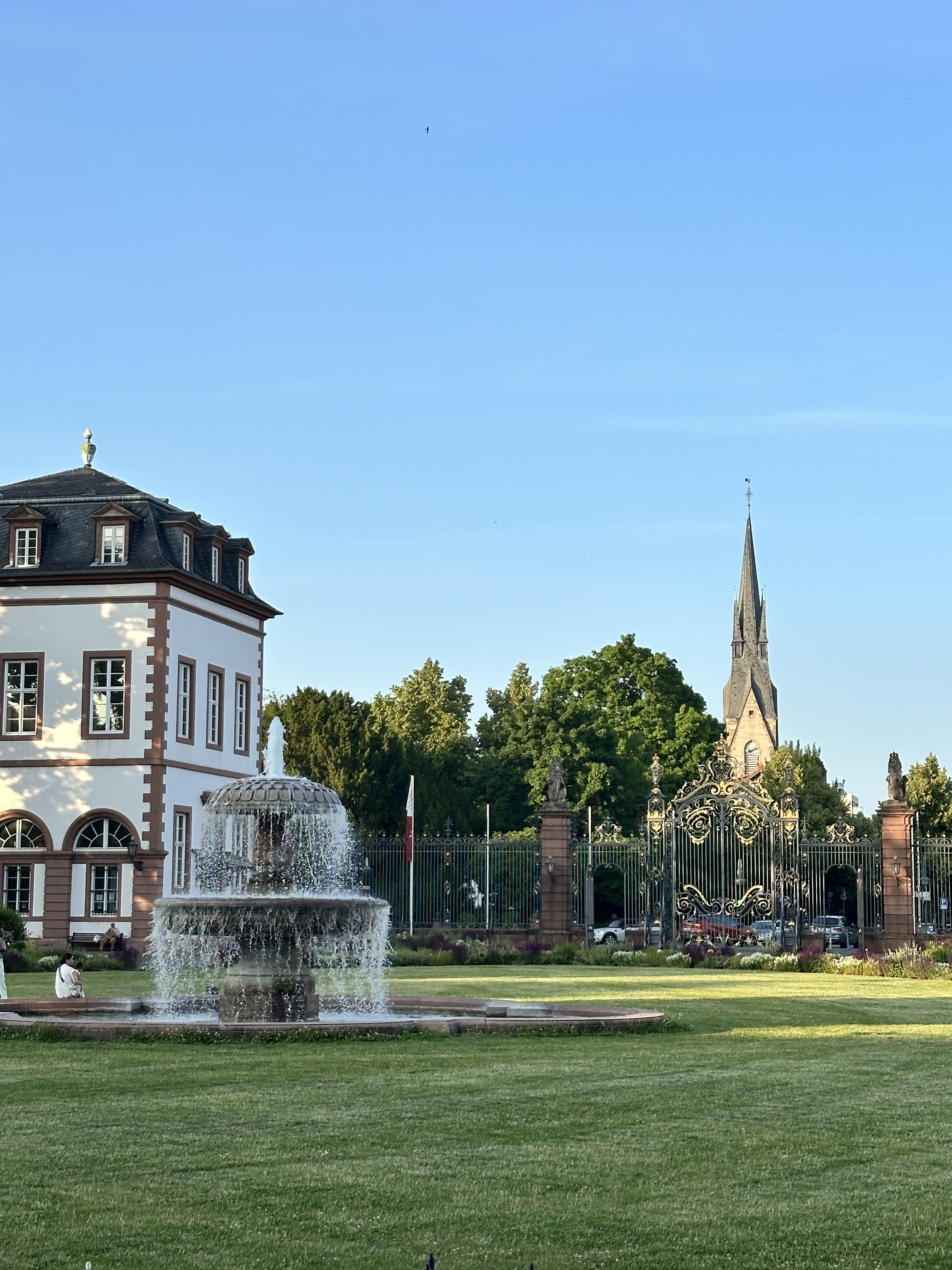 A serene park scene featuring a cascading fountain in the foreground, a grand historic building to the left, ornate iron gates in the background, and a church steeple rising above lush green trees under a clear blue sky.