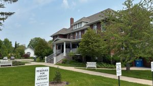 A stately old brock building with a large porch, trees and a lawn un front. In the foreground are some signs that say "Library parking only"