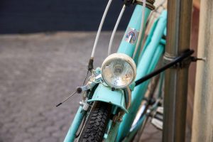 A close-up view of a vintage turquoise bicycle, featuring a front headlight mounted on the fork near the wheel. The bike’s metallic structure and tire tread are visible, with the headlight showing some wear.
