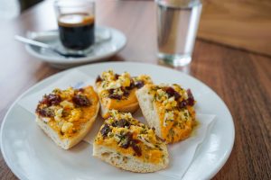 A plate of four open-faced sandwiches on a white napkin. Each sandwich has a thick layer of salmorejo topped with chopped herbs, small pieces of boiled egg, and bits of ibérico. There are a small glass of water and a cup of dark coffee in a saucer are visible in the background.
