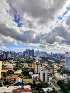 A dramatic view of Cebu City, Philippines, showcasing a mix of low-rise neighborhoods and modern high-rises under a sky filled with thick, textured clouds. The scene captures the vibrant contrast of urban life and tropical weather.
