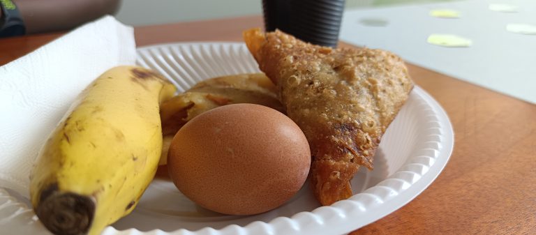 A paper plate holds a banana, a brown boiled egg, a fried triangular pastry, and a slice of potato, arranged alongside a paper towel. In the background, there is a dark cup and a partially visible table.