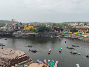 A scenic view of a river surrounded by rocky cliffs, with multiple boats floating on the water. In the background, there are buildings, including a bright yellow structure, and a suspension bridge. Colorful canopies and bustling activity along the riverbank indicate a lively market area. The sky is overcast, adding a soft light to the scene.