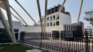 A modern building with a white facade and large glass windows is surrounded by a black wrought-iron fence. 