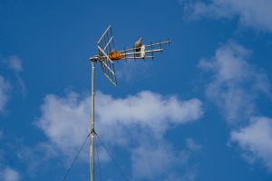 A dove is perched on an antenna mounted on a tall pole against a bright blue sky with scattered white clouds. The antenna features several horizontal and vertical metal elements, and the dove appears to be surveying the area.