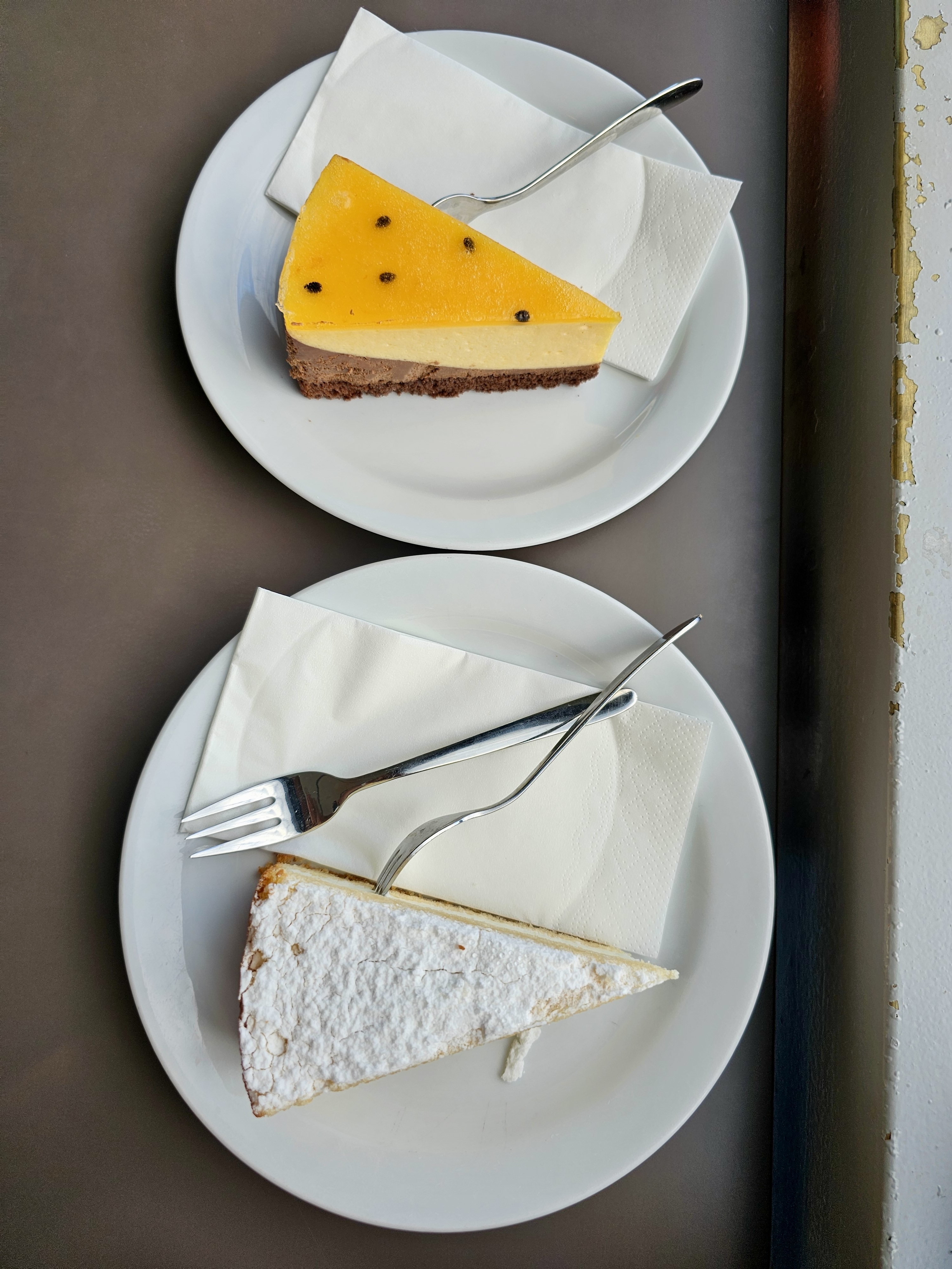 Two plates, each holding a slice of cake and a fork. The top plate has a slice of passion fruit cake with a multi-layered base, while the bottom plate contains a slice of vanilla meringue cake dusted with powdered sugar. Captured from Danube tower, Vienna.