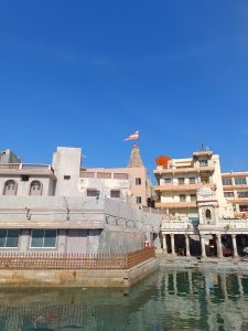 A view of a riverbank with buildings, including a temple featuring a decorative spire and a flag atop it. 
