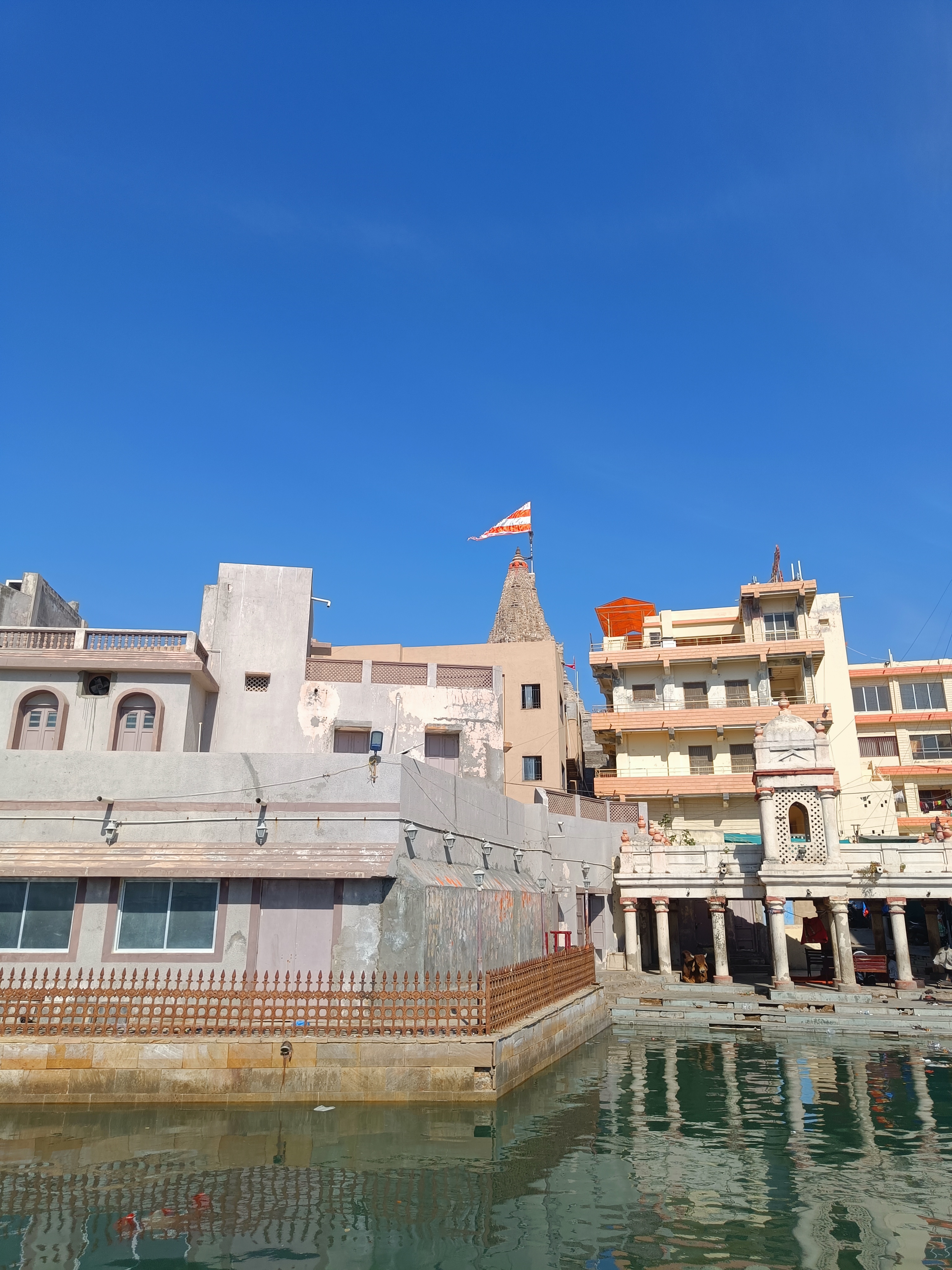 A view of a riverbank with buildings, including a temple featuring a decorative spire and a flag atop it.