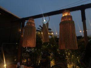 An outdoor dining area at dusk features three large, glowing, straw-like hanging lamps suspended from a wooden beam. Warm string lights twinkle in the background. Surrounded by lush greenery, the cozy setup includes a few chairs and tables, creating an inviting, tranquil atmosphere.
