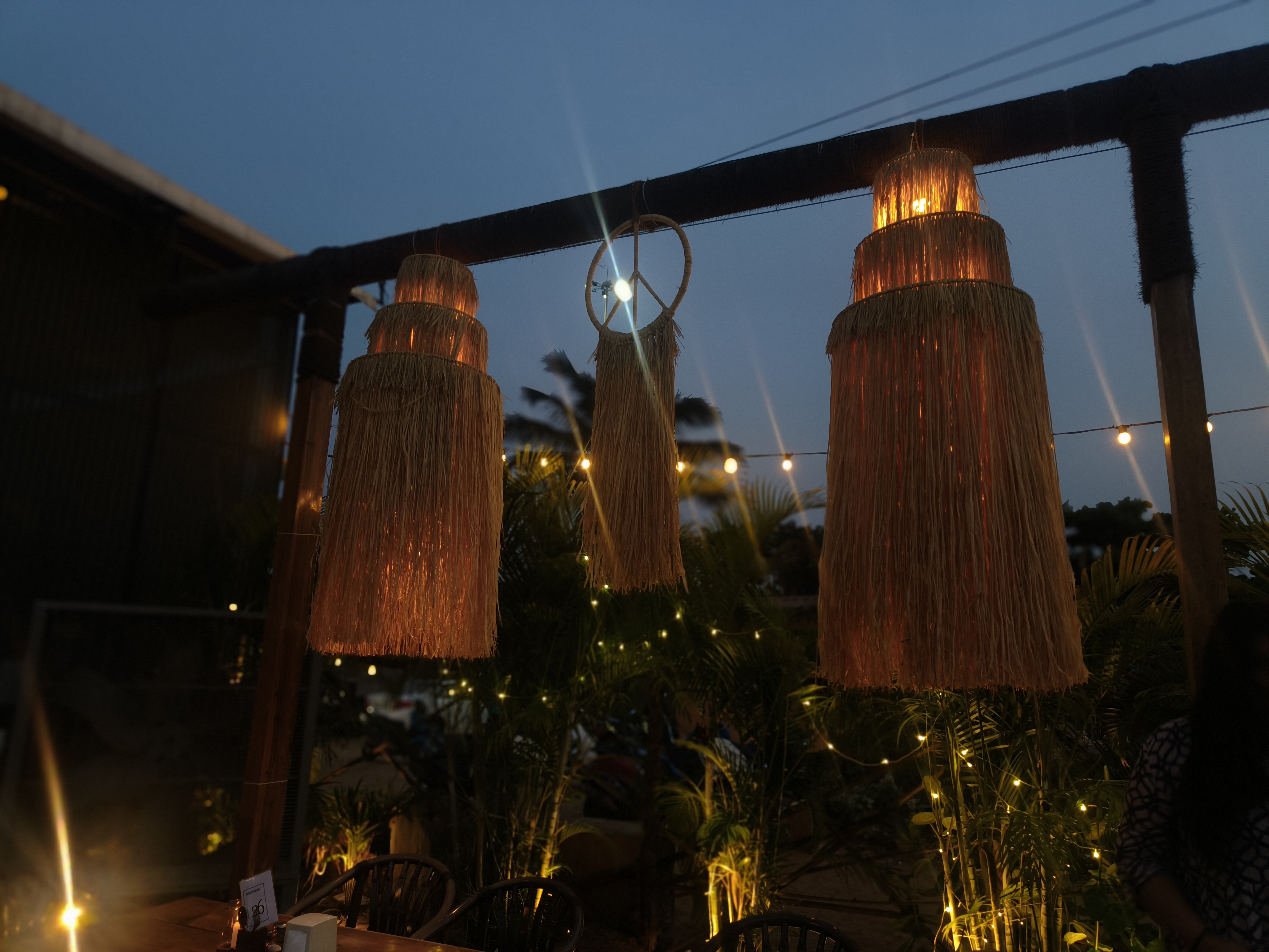 An outdoor dining area at dusk features three large, glowing, straw-like hanging lamps suspended from a wooden beam. Warm string lights twinkle in the background. Surrounded by lush greenery, the cozy setup includes a few chairs and tables, creating an inviting, tranquil atmosphere.