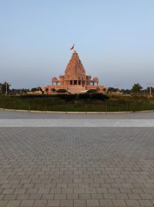 A large, intricately designed  Hindu temple stands majestically in the background, showcasing ornate architectural features and a prominent golden dome topped with a flag. In the foreground, there is a wide expanse of a tiled walkway
