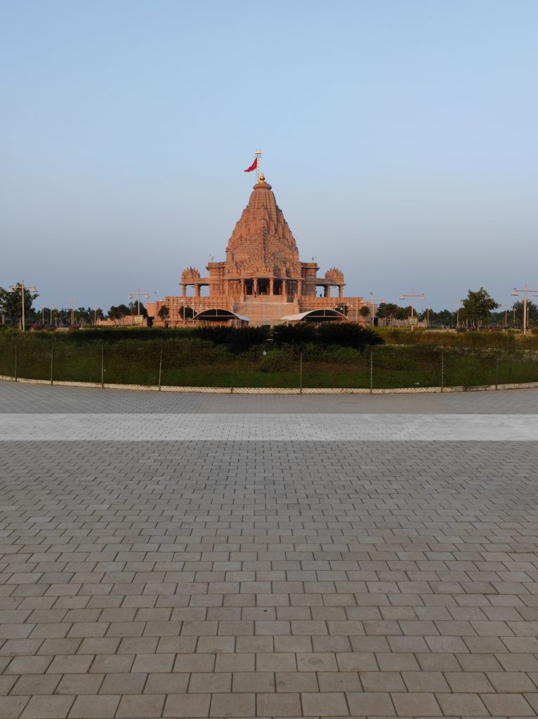 A large, intricately designed  Hindu temple stands majestically in the background, showcasing ornate architectural features and a prominent golden dome topped with a flag. In the foreground, there is a wide expanse of a tiled walkway