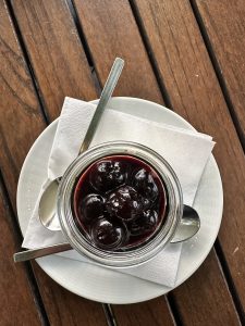 A glass jar filled with glossy, dark cherries in syrup sits on a white plate with a paper napkin and two spoons, ready to be shared as a sweet treat.