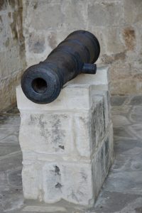 Old black iron cannon on a stone pedestal at Limassol Castle, Cyprus, with a weathered stone wall behind.