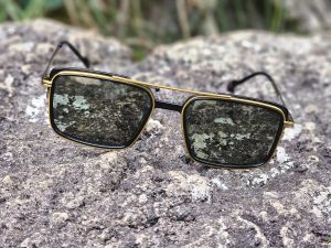 Black and gold eyeglasses with tinted lenses rest on a rough rock at Needle Rock, Gudalur, with blurred plants in the background.
