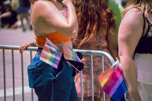 People stand near a metal barricade at a Pride event. One person wears an orange crochet top and jeans with two pastel Progress Pride flags tucked into their waistband. Another person nearby has a small Progress Pride flag clipped to their skirt.