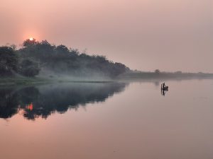 A serene landscape featuring a calm river reflecting the soft colors of a sunset. On the left side, a solitary figure is paddling a small boat, while gentle mist rises from the water