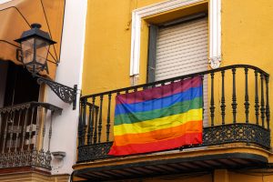 A vibrant rainbow flag hangs from a black wrought iron balcony, set against a bright yellow wall. The balcony features intricate detailing, and a decorative lantern is mounted on the wall nearby.