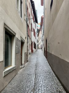 Narrow cobblestone street lined with tall, closely packed buildings featuring red and white facades, seen on a cloudy day.
