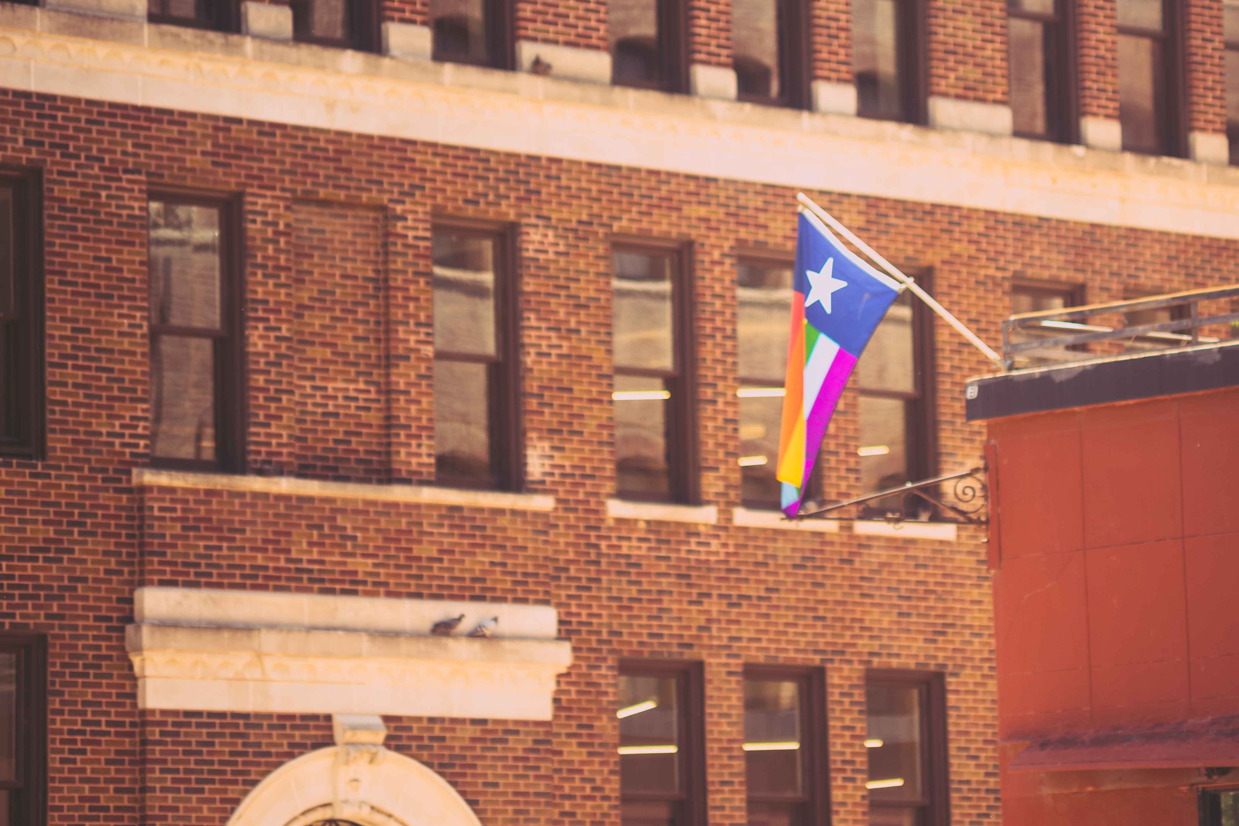 A Flag hangs on a diagnal from the side of one building. The flag is styled after the Texas flag with a lone star but instead of the two stripes it features multiple rainbow stripes. The background is a brick building.