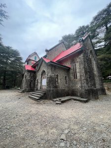 A historic stone church with red roofs is surrounded by tall trees.
