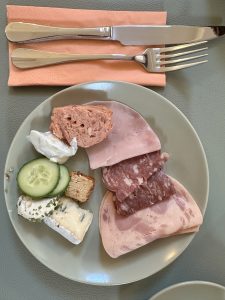 A white plate full of pork slices (cutlets), cucumber slices, butter, etc on the table. Fork and Knife are also on the table of a restaurant.  