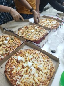 Multiple open pizza boxes with various topped pizzas on a table while people reach for slices during a casual meal.
