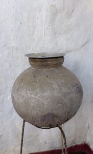 A large, round, weathered clay pot sits on a metal stand. The pot has a smooth, slightly cracked surface and a small lip at the top, which is covered by a shiny, circular metal lid. 