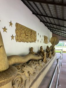 A long, intricately carved relief sculpture featuring figures of children and a serpent, depicted in a flowing wave-like pattern. The figures are shown actively engaging with the serpent, with some holding onto it.