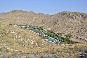 A campsite or small village is set in a dry valley, surrounded by rocky hills. The land is brown with little plants, and the sky is clear and blue. Taken in Almería, Spain.