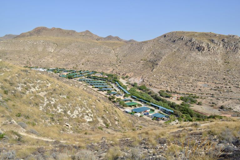 A campsite or small village is set in a dry valley, surrounded by rocky hills. The land is brown with little plants, and the sky is clear and blue. Taken in Almería, Spain.