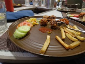 A brown plate with cucumber and tomato slices, corn salad, French fries, Manchurian balls, and red sauce, with a fork placed on the side.
