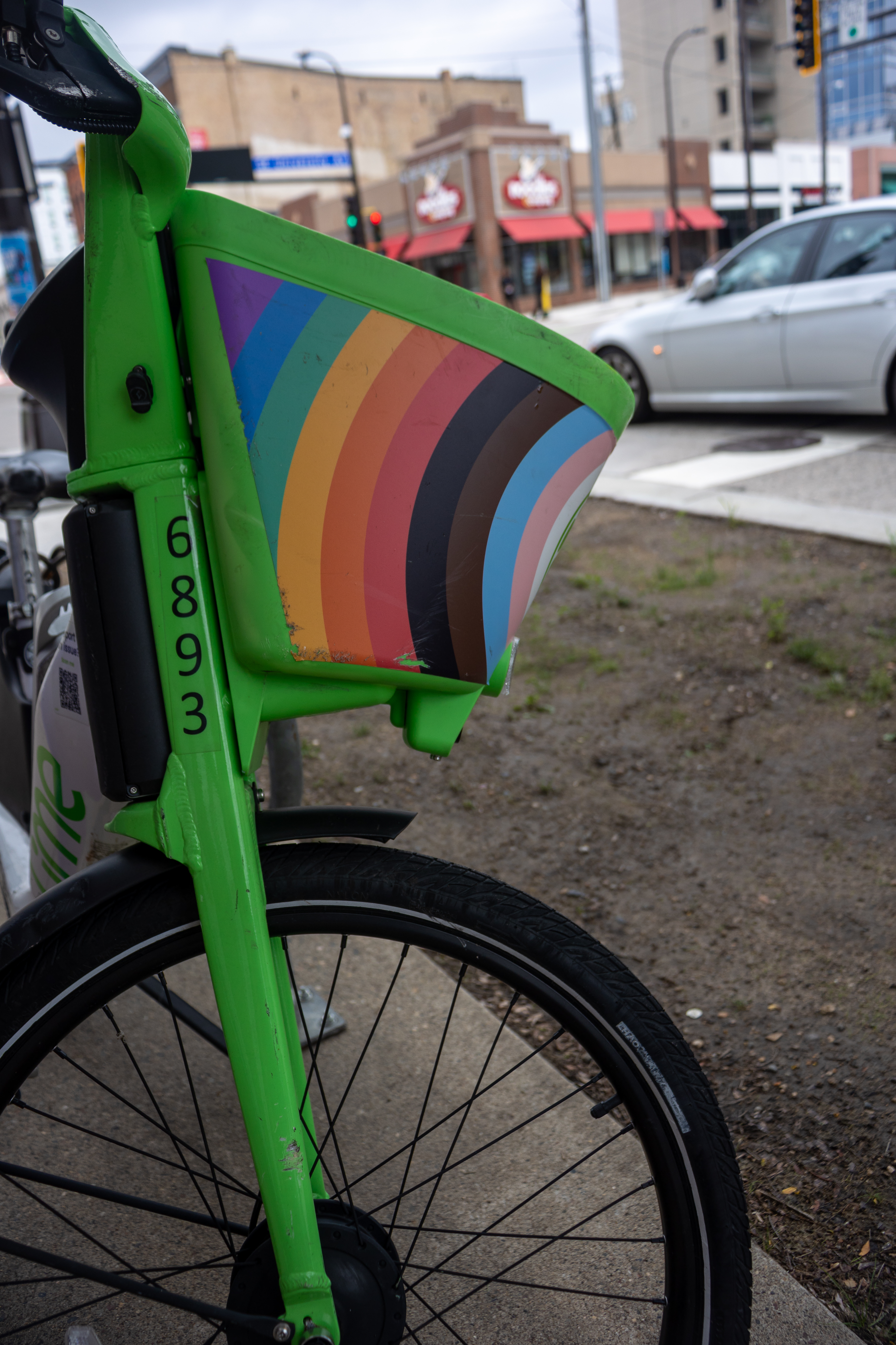 The front of a green bicycle with a rainbow pride progress flag painted on the basket. In the background is a city street with a car and shops