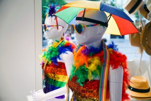 Two mannequins dressed in vibrant, colorful outfits are displayed in a shop window. Each mannequin wears a sparkly, sequined top in rainbow colors, adorned with fluffy feather boas in various bright shades. They also sport round, rainbow-striped sunglasses and small, colorful umbrellas hatting their heads.